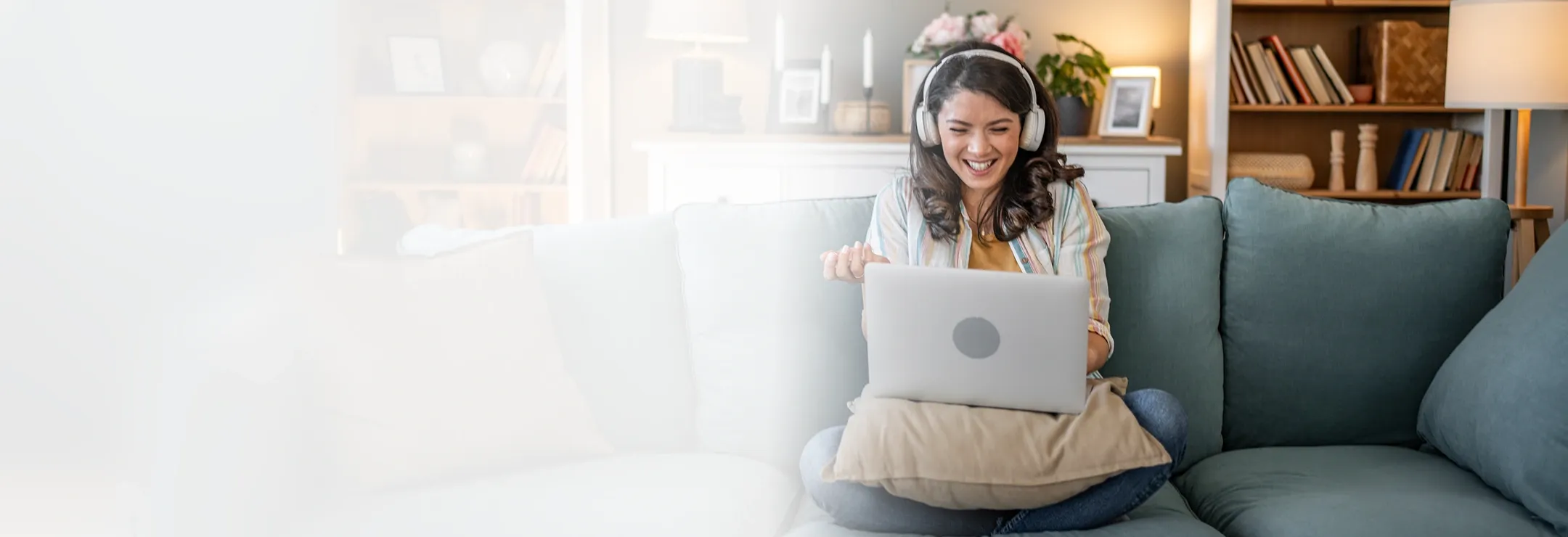 A smiling woman wearing headphones sits cross-legged on a couch with a laptop in front of her, resting on a cushion.